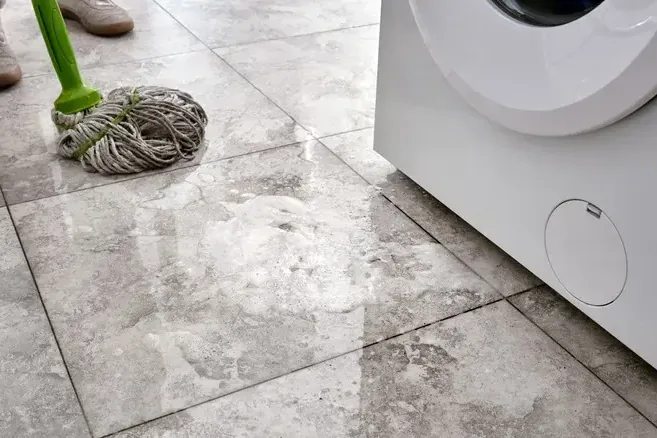 Wet tile floor with a mop and bucket beside a front-load washer after a leak